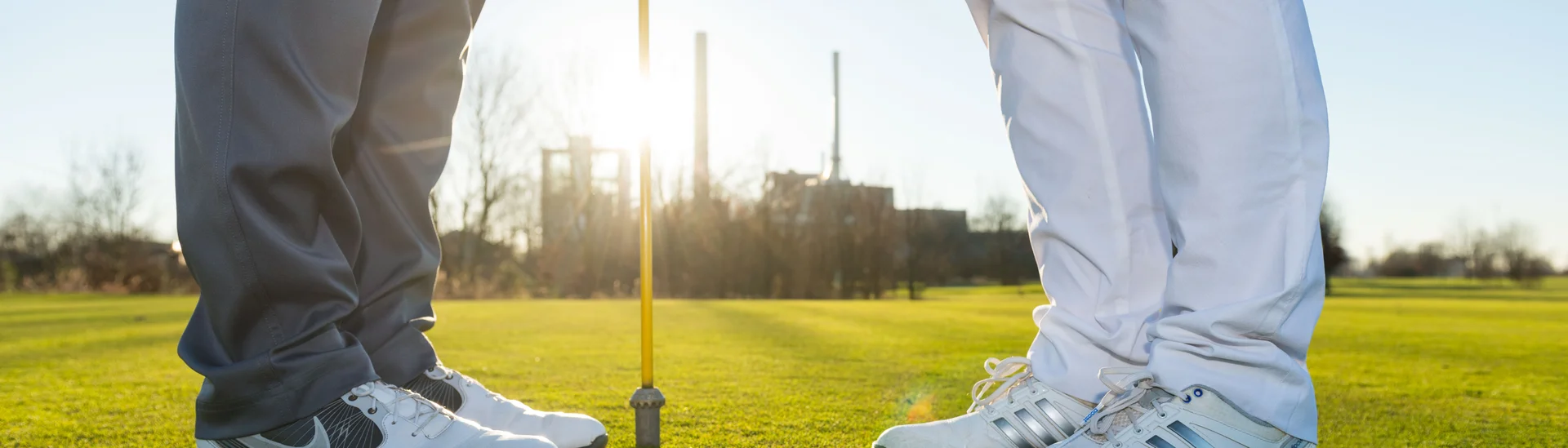zwei Personen in Turnschuhen stehen voreinander vor einem Golfloch mit Fahne, Ausschnitt bis kurz über dem Knie